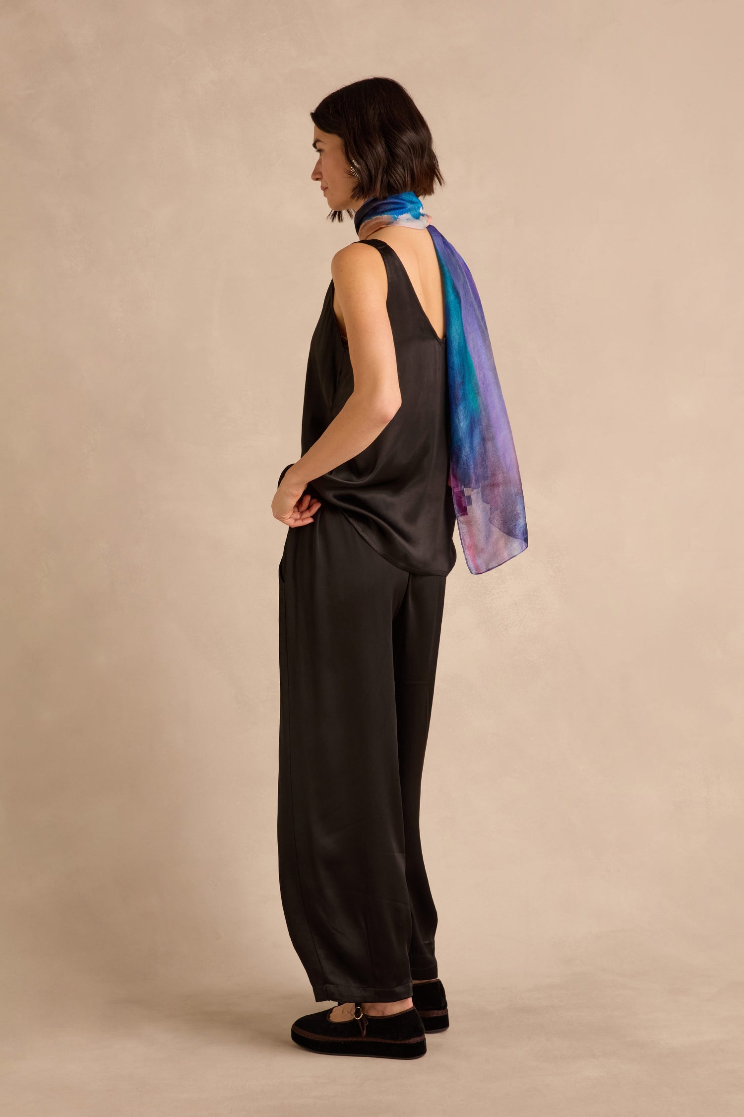 A woman stands in profile against a beige background, wearing a black sleeveless top, Satin Back Crepe Bubble Trousers in black, black shoes, and a blue-purple patterned scarf around her neck.