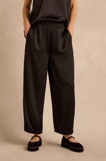 Wearing the Satin Back Crepe Bubble Trousers, a person stands with hands in pockets against a plain beige background, paired with a black sleeveless top and black shoes.