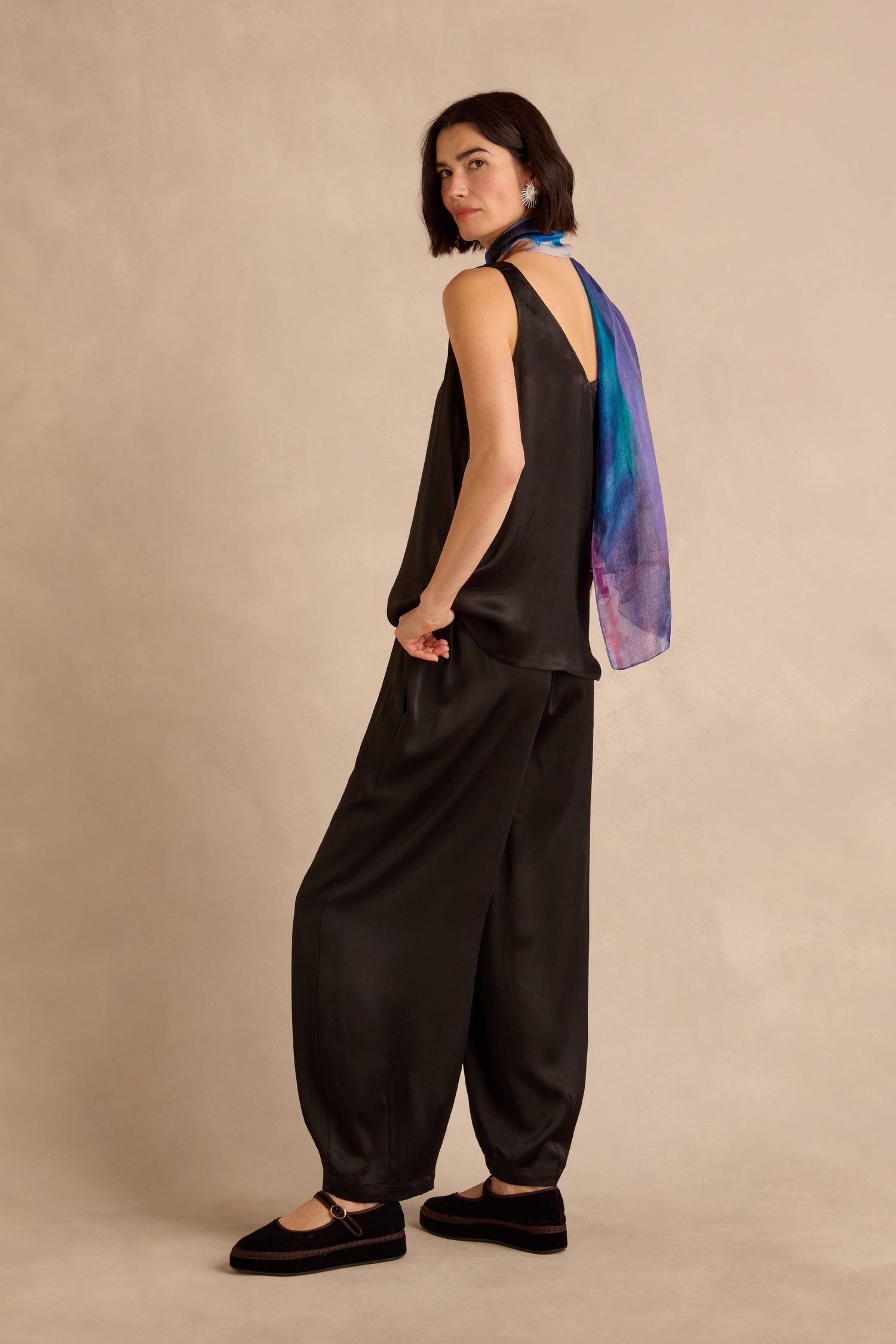 A woman stands against a beige background wearing a loose black sleeveless jumpsuit paired with Satin Back Crepe Bubble Trousers, black shoes, and a blue and purple scarf.