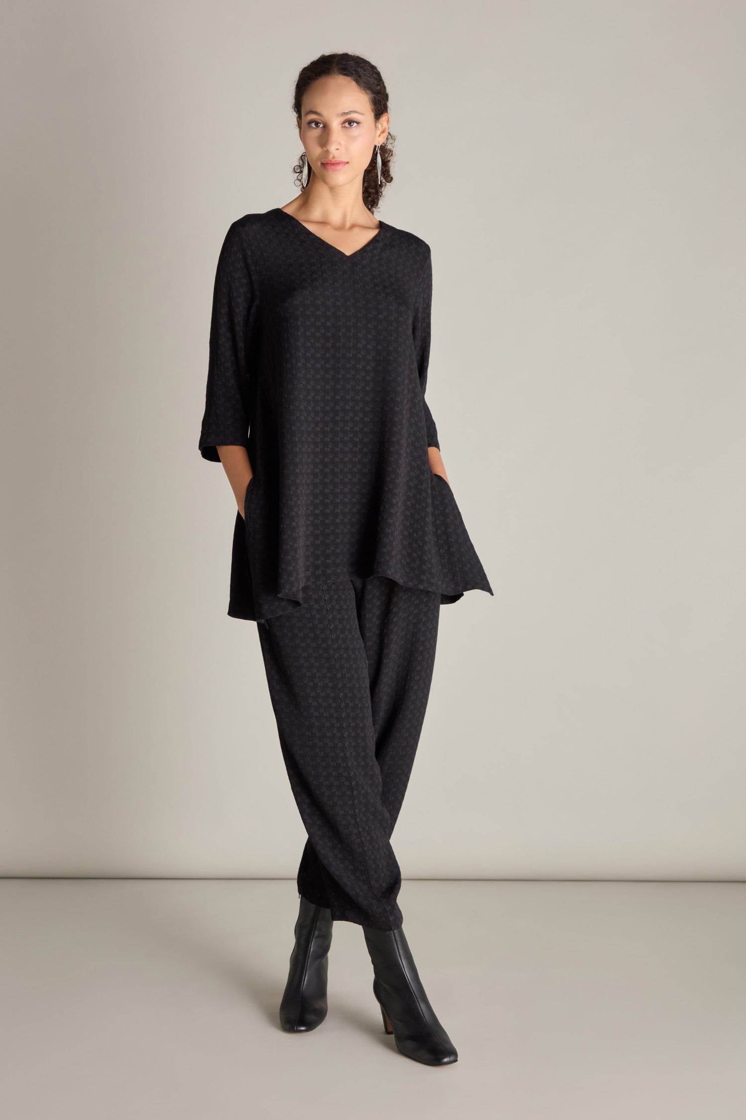 A woman stands against a plain background wearing a Small Square Jacquard Tunic with 3/4 length sleeves and matching pants. The tunic features a V-neckline, which is complemented by black ankle boots.