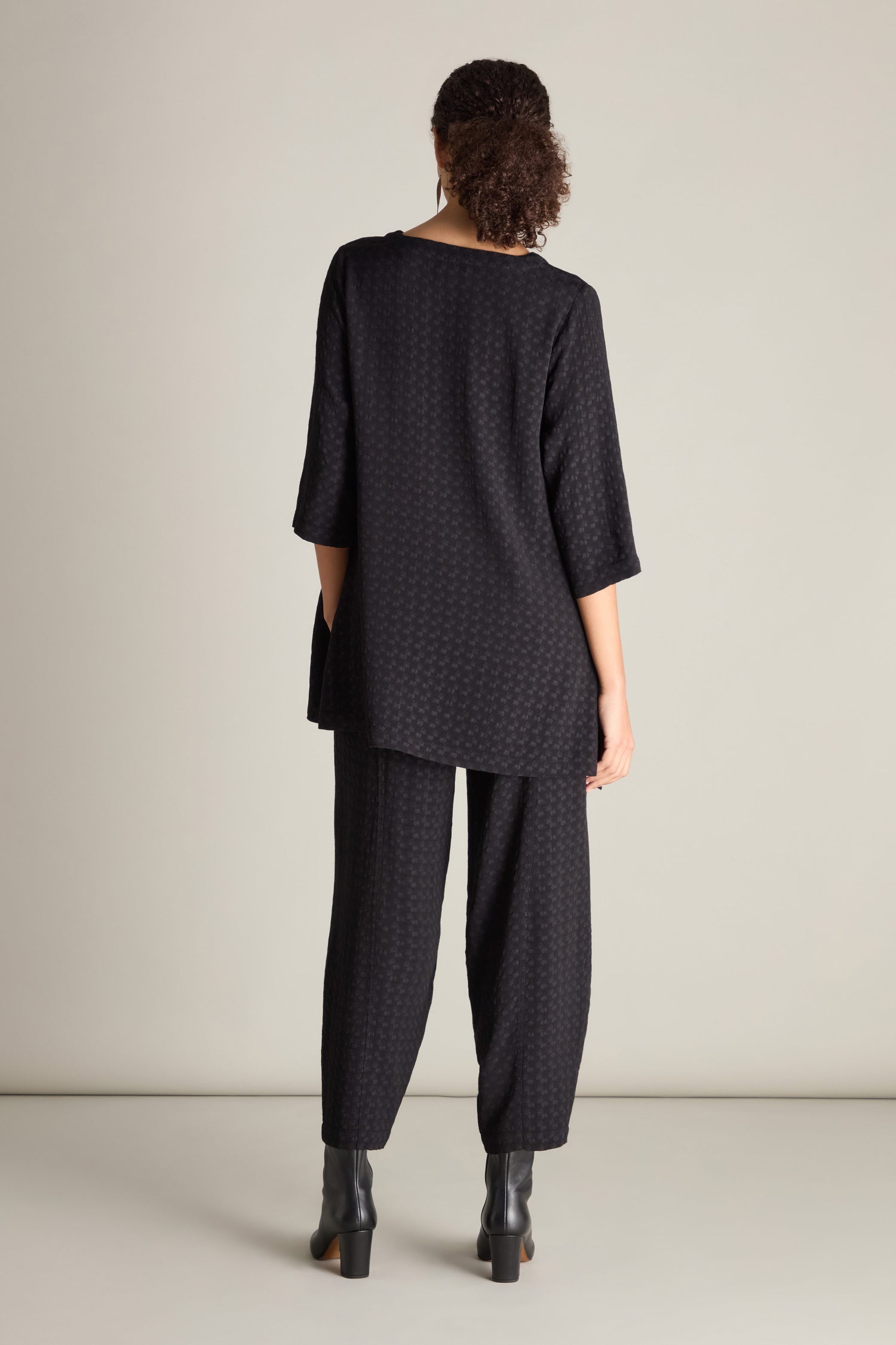 Person with curly hair, wearing the Small Square Jacquard Tunic, which boasts a black patterned design and an A-line silhouette, paired with matching pants. The tunic features a V-neckline. The person is standing in a room with a light-colored wall, facing away from the camera.