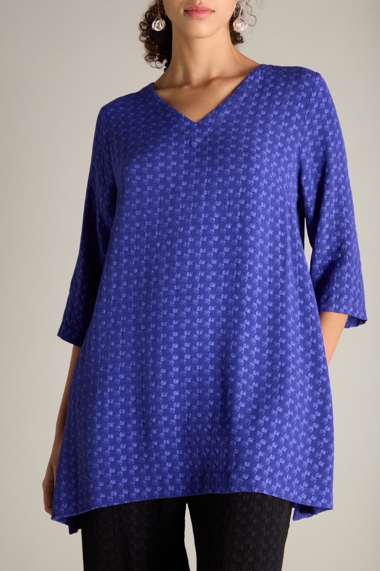 Woman wearing a blue Small Square Jacquard Tunic with a V-neckline and three-quarter sleeves, paired with black pants.