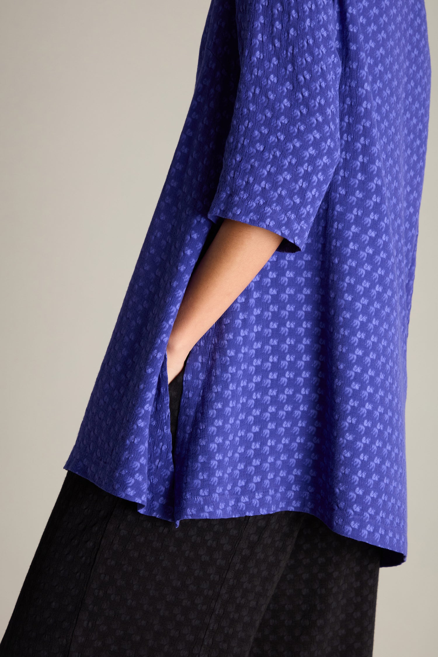 A person wearing the Small Square Jacquard Tunic featuring a V-neckline, 3/4 length sleeves, and side slits stands with one hand in their pocket, paired with dark trousers.