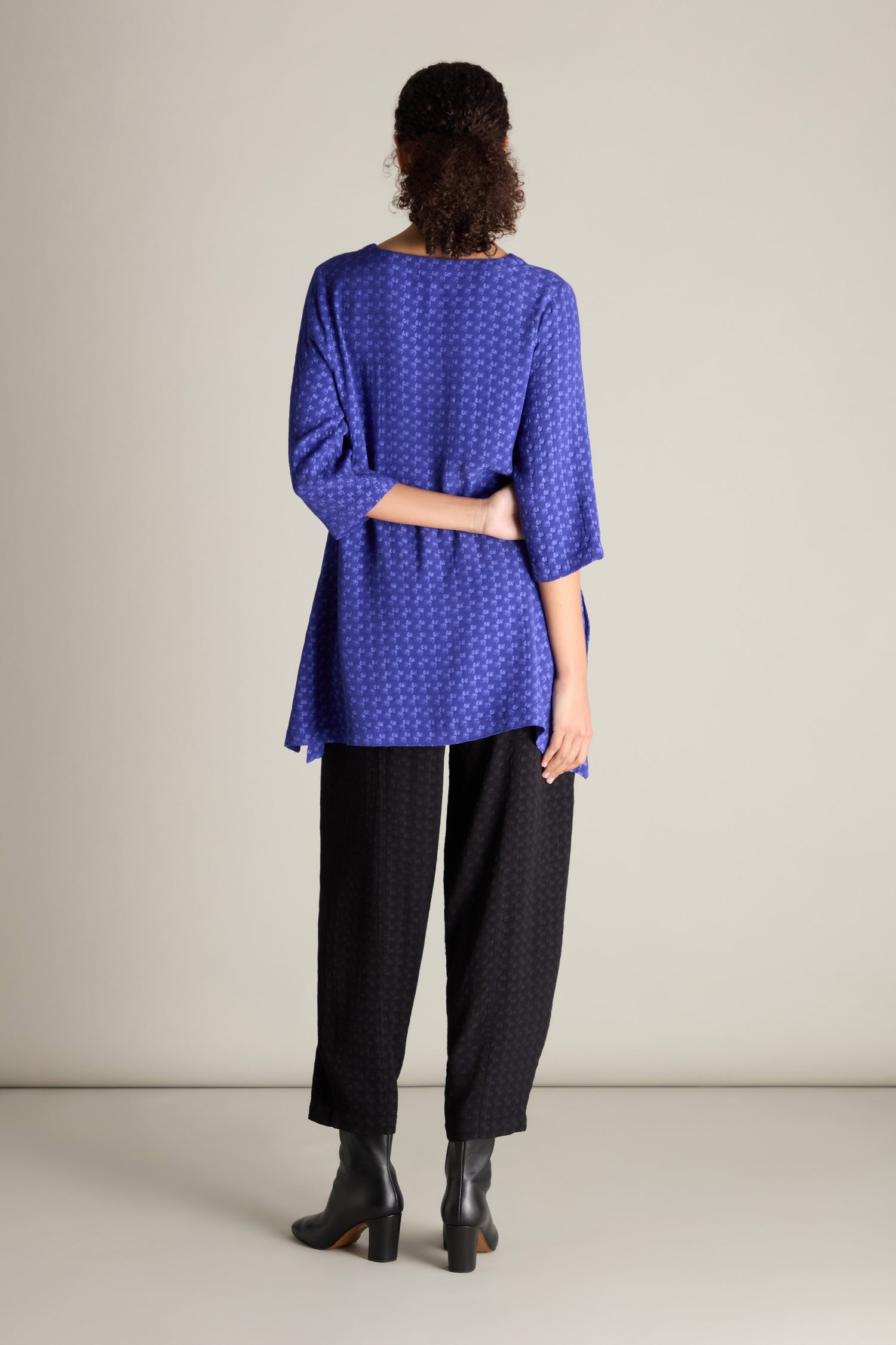 A person is standing with their back to the camera, dressed in a Small Square Jacquard Tunic with a V-neckline and 3/4 length sleeves, paired with black pants and black heeled boots. Their left arm is resting behind their back.