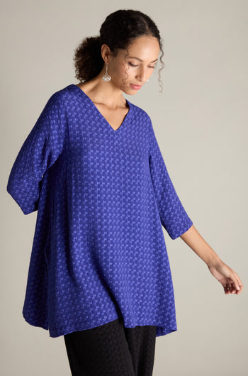 A person dressed in a Small Square Jacquard Tunic featuring a V-neckline and three-quarter length sleeves looks down while posing against a simple backdrop.