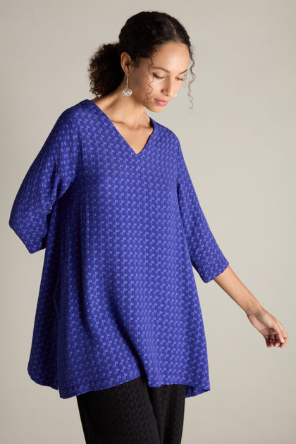 A person dressed in a Small Square Jacquard Tunic featuring a V-neckline and three-quarter length sleeves looks down while posing against a simple backdrop.