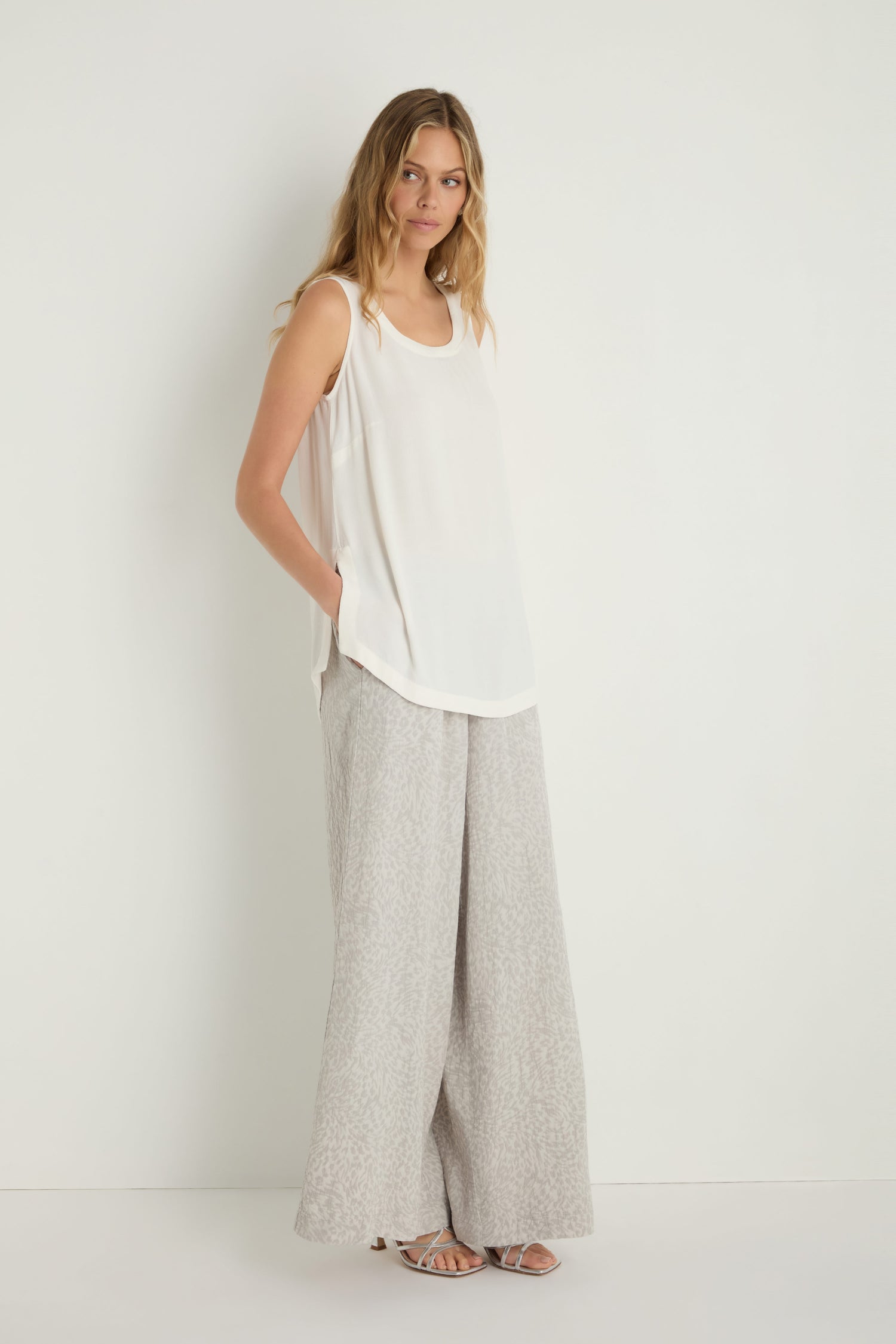 Woman stands against a white background wearing a loose white sleeveless top, wide-leg light gray pants, sandals, and the Crepe Vest—a perfect effortless lightweight layering piece—with hands in her pockets.