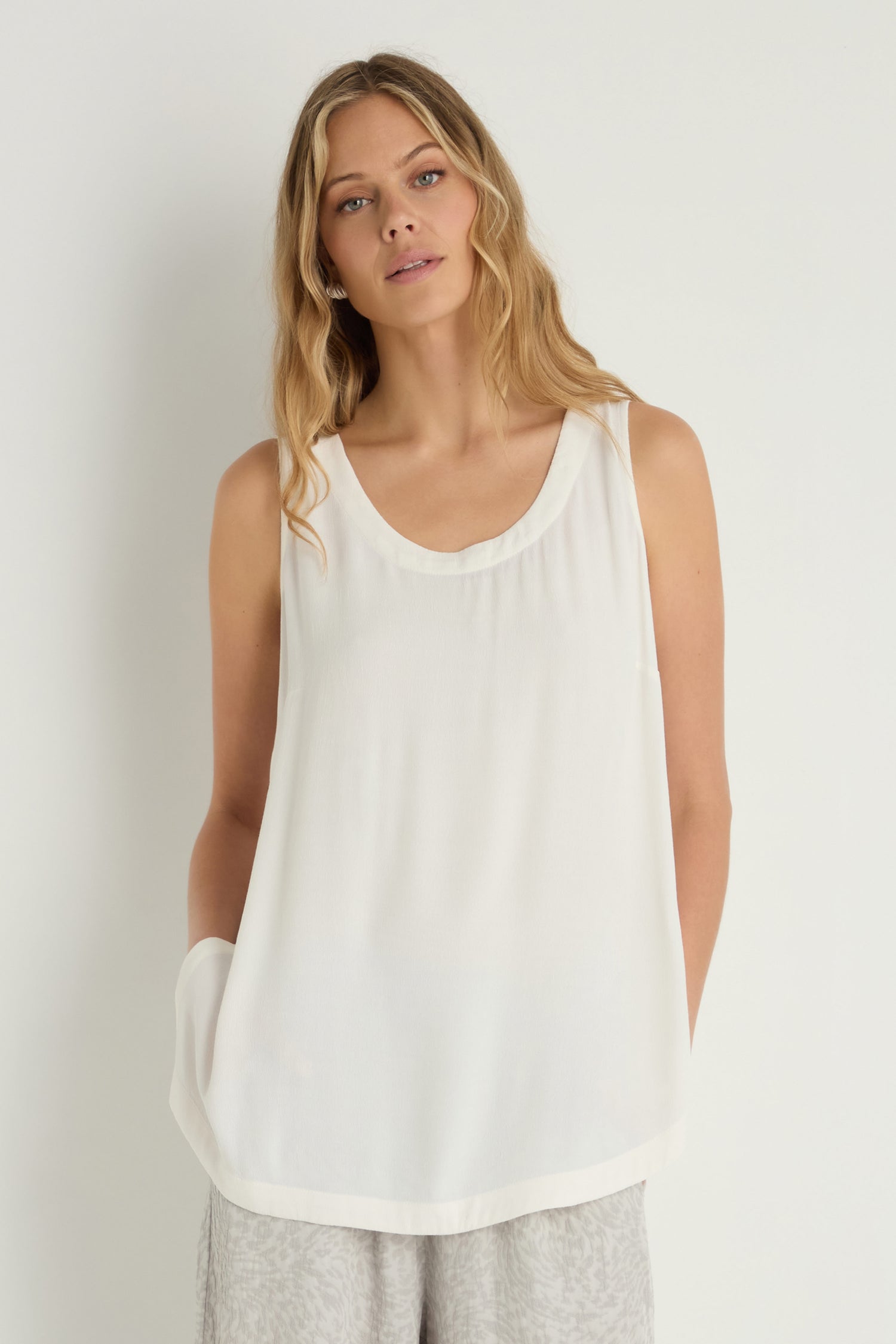 A woman stands against a plain background wearing a sleeveless white Crepe Vest made from viscose crepe and light-colored pants, hands in her pockets—an ideal lightweight layering piece.