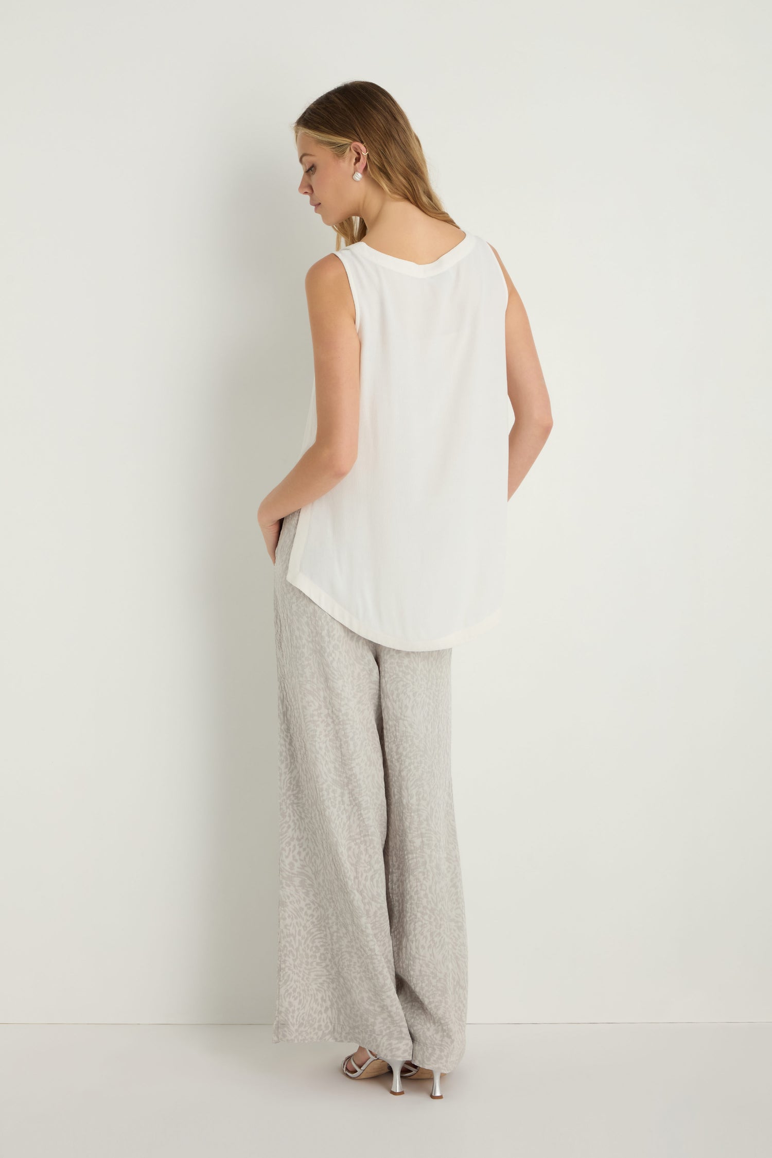 A woman stands against a white wall, facing away. She wears a sleeveless white Crepe Vest, light gray wide-leg viscose crepe pants, and heeled sandals. Her hands are tucked casually into her pockets.