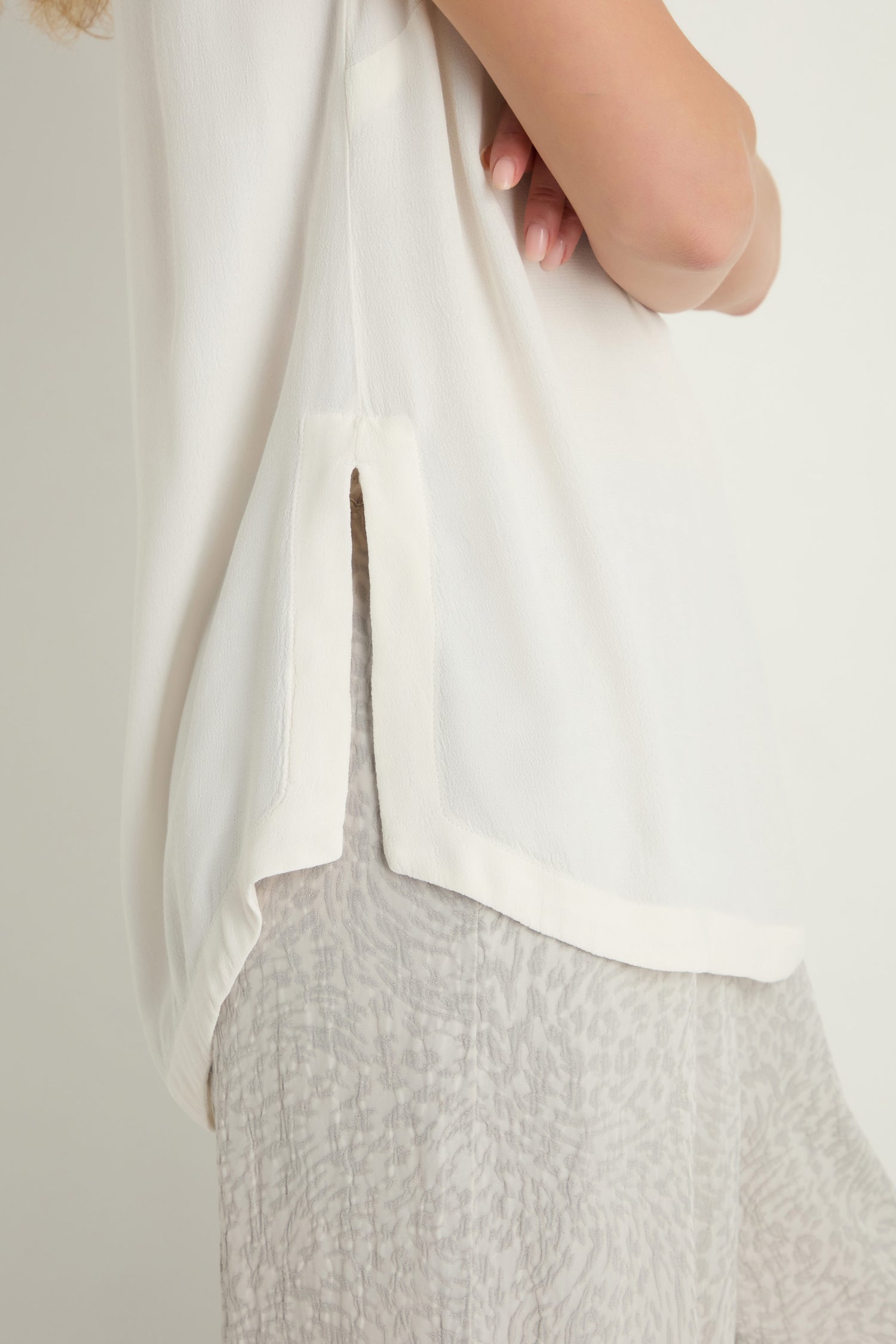 A person in a white Crepe Vest, a lightweight layering piece crafted from viscose crepe, paired with light patterned pants, is shown from shoulder to mid-thigh with arms crossed.