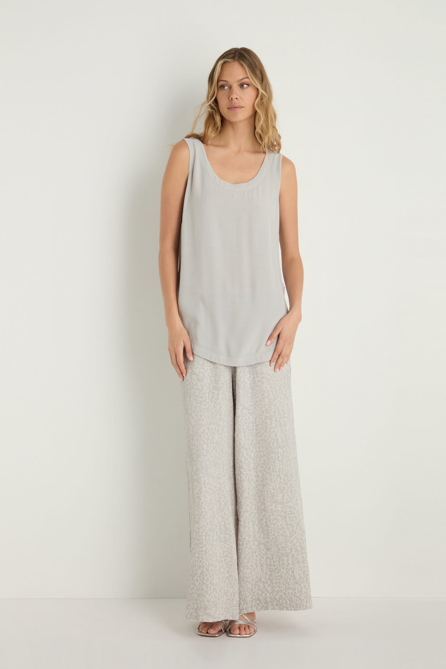 A woman stands against a white wall in a sleeveless gray top, wide-leg patterned pants, and sandals, effortlessly styling her look with a Crepe Vest—a lightweight layering piece in soft viscose crepe.