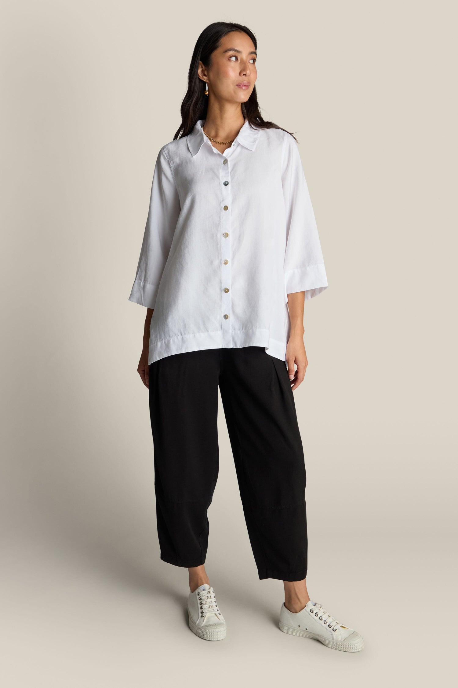 A woman wearing a Soft Tencel Flared Hem Shirt with mother-of-pearl buttons and black wide-leg pants stands against a beige background. She is looking off to the side with a neutral expression, showcasing a flattering silhouette.