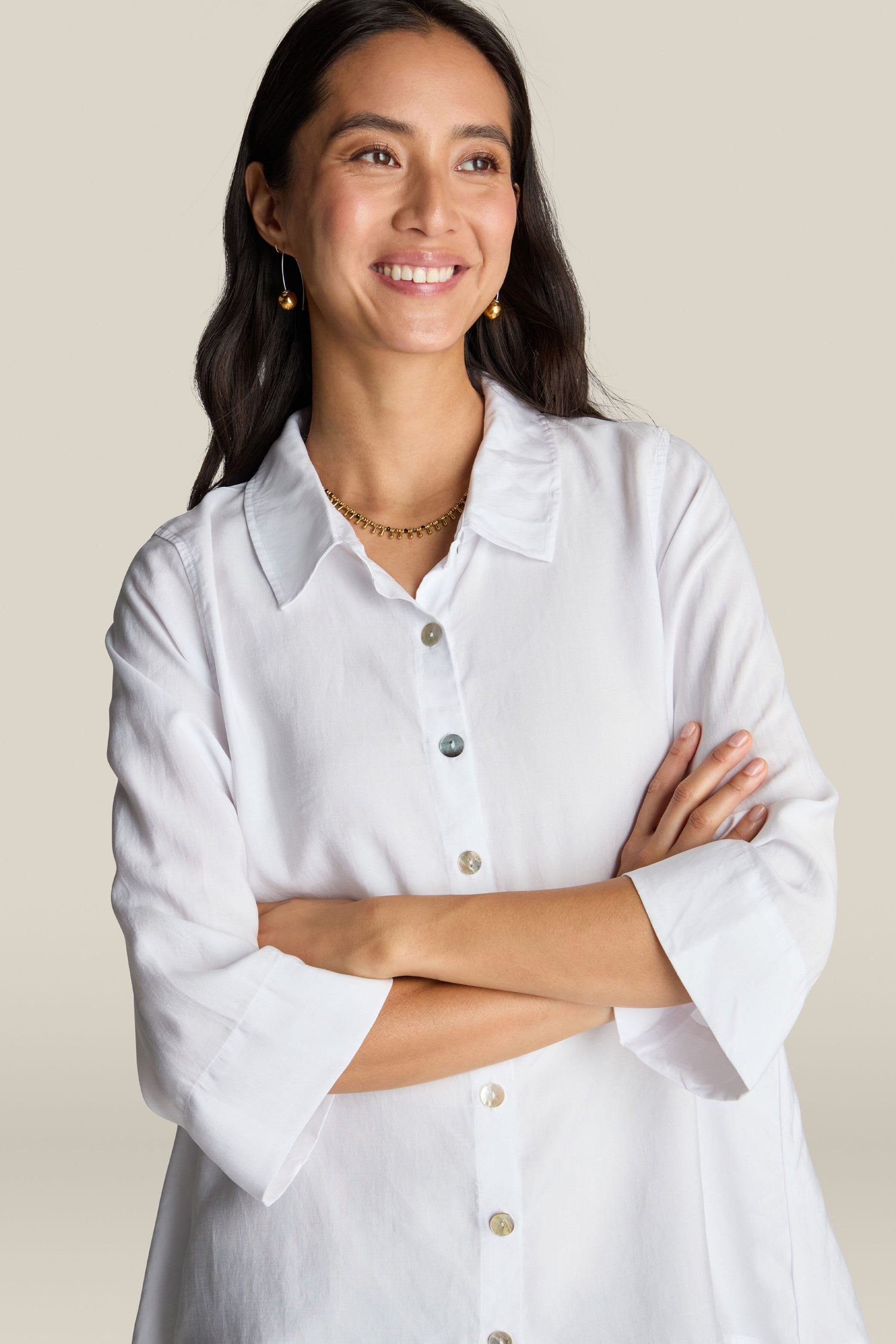 A woman with long dark hair, wearing a Soft Tencel Flared Hem Shirt with mother-of-pearl buttons and earrings, smiles with her arms crossed in front of a neutral background.