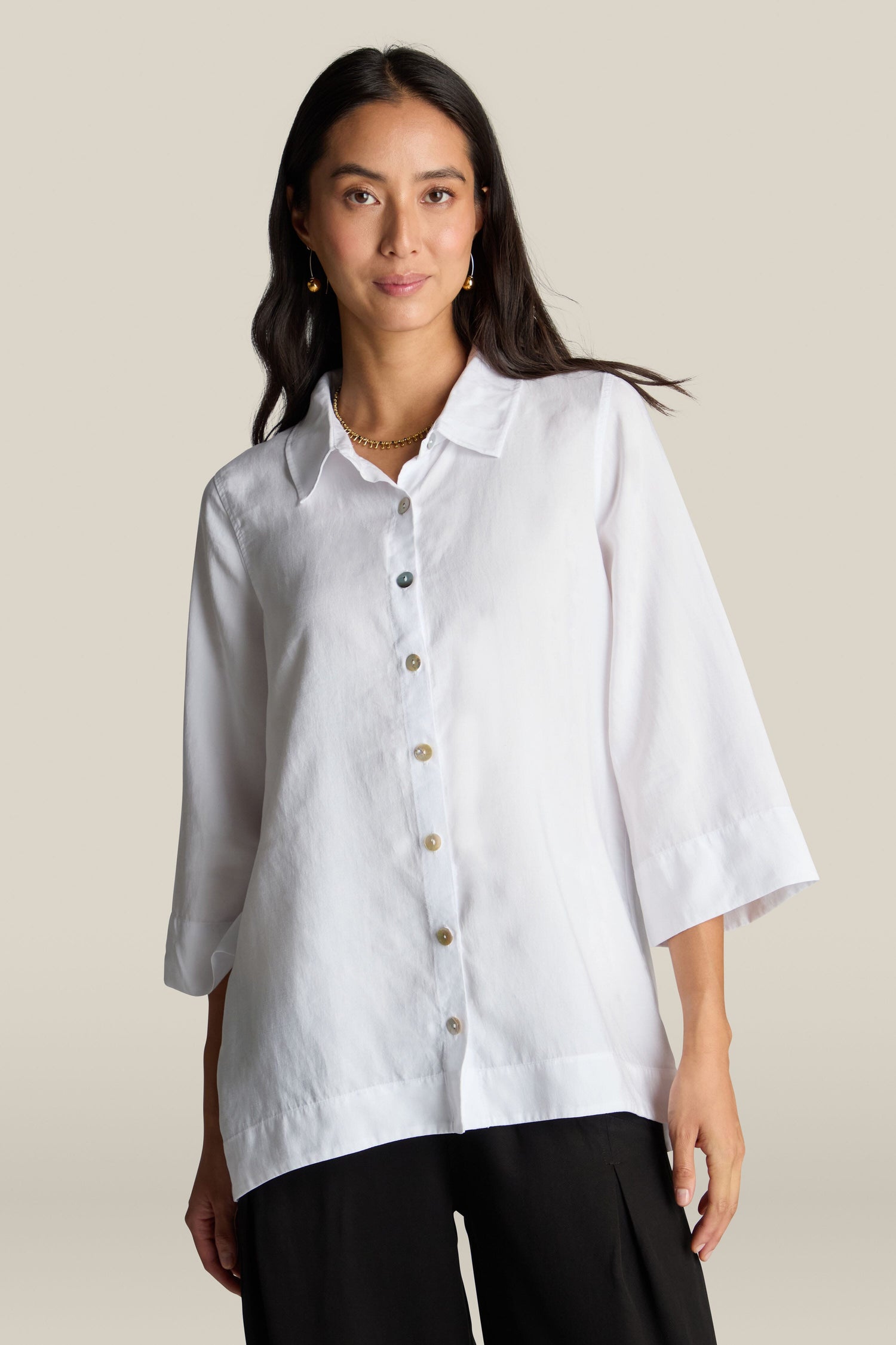 A person with long dark hair wears a Soft Tencel Flared Hem Shirt with mother-of-pearl buttons and black pants, standing against a light beige background.