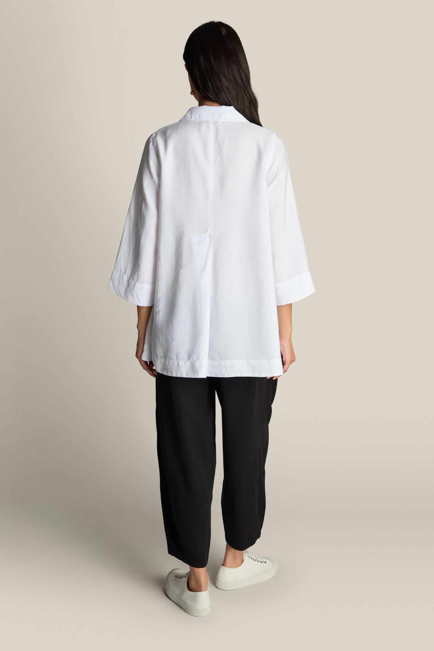 A person with long dark hair, facing away from the camera, is wearing a white Soft Tencel Flared Hem Shirt with mother-of-pearl buttons, black pants, and white sneakers against a plain beige background.