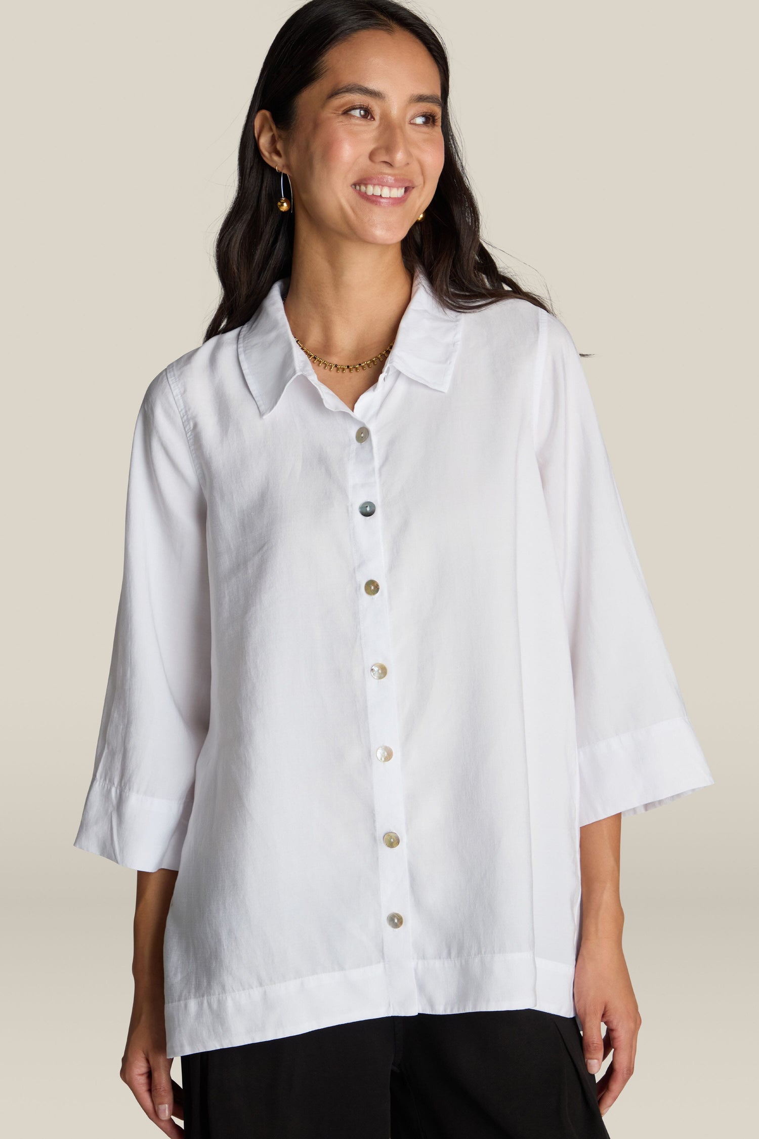 A woman standing, wearing a Soft Tencel Flared Hem Shirt with mother-of-pearl buttons and black pants. She is smiling and looking slightly to the side, showcasing a flattering silhouette. The background is plain beige.