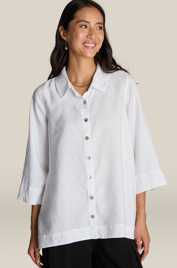 A woman standing, wearing a Soft Tencel Flared Hem Shirt with mother-of-pearl buttons and black pants. She is smiling and looking slightly to the side, showcasing a flattering silhouette. The background is plain beige.