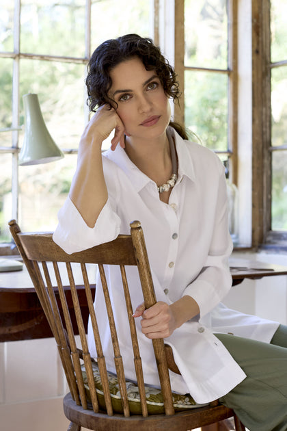 A woman with curly hair, wearing a white Soft Tencel Flared Hem Shirt adorned with mother-of-pearl shell buttons and green pants, sits on a wooden chair by a window, resting her head on her hand.