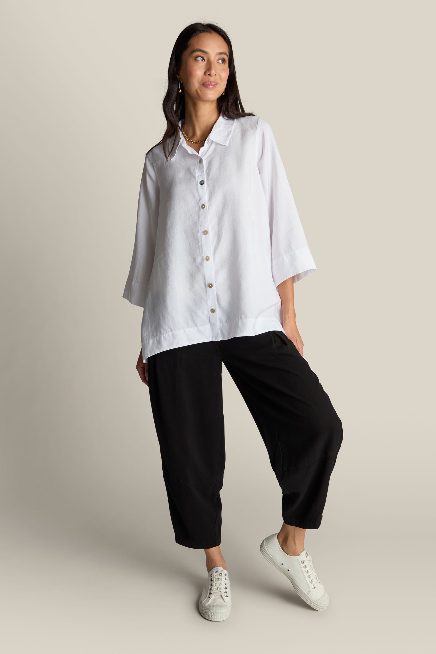 A woman stands against a plain background wearing the Soft Tencel Flared Hem Shirt with mother-of-pearl buttons, paired with black pants and white sneakers. She is smiling, her hands relaxed by her sides.