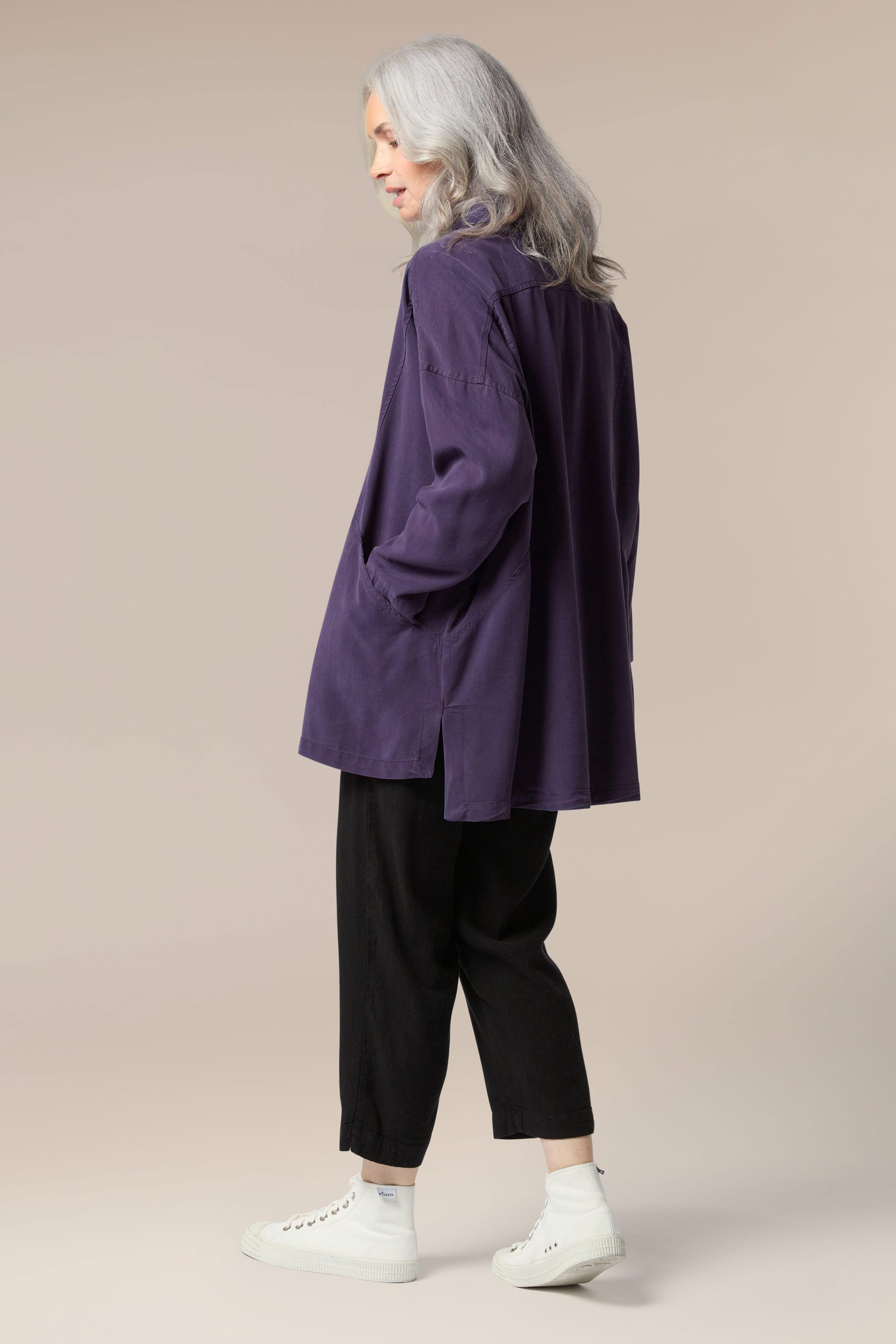 A model is wearing a purple twill shirt open with a collar, button front and pockets. It is paired with a white top underneath, black trouser, and white trainers.