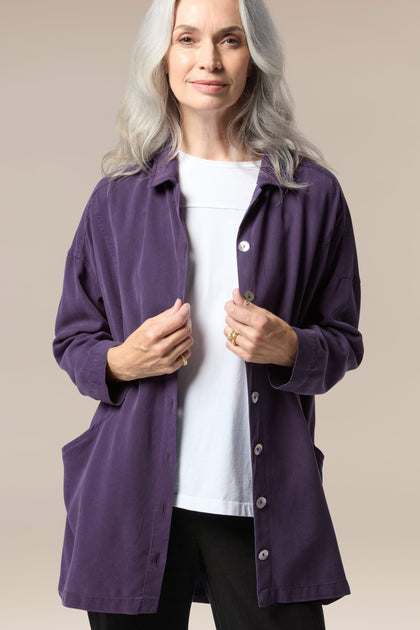 A model is wearing a purple twill shirt open with a collar, button front and pockets. It is paired with a white top underneath and black trousers.