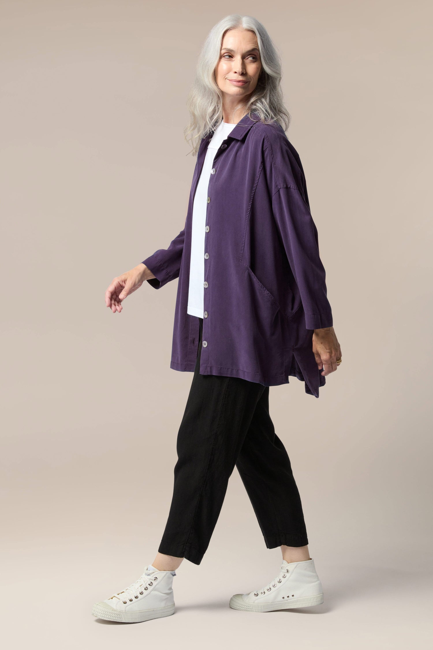 A model is wearing a purple twill shirt open with a collar, button front and pockets. It is paired with a white top underneath, black trouser, and white trainers.