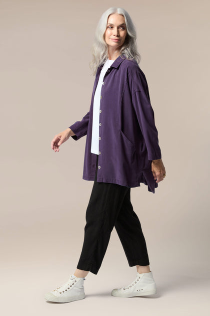 A model is wearing a purple twill shirt open with a collar, button front and pockets. It is paired with a white top underneath, black trouser, and white trainers.