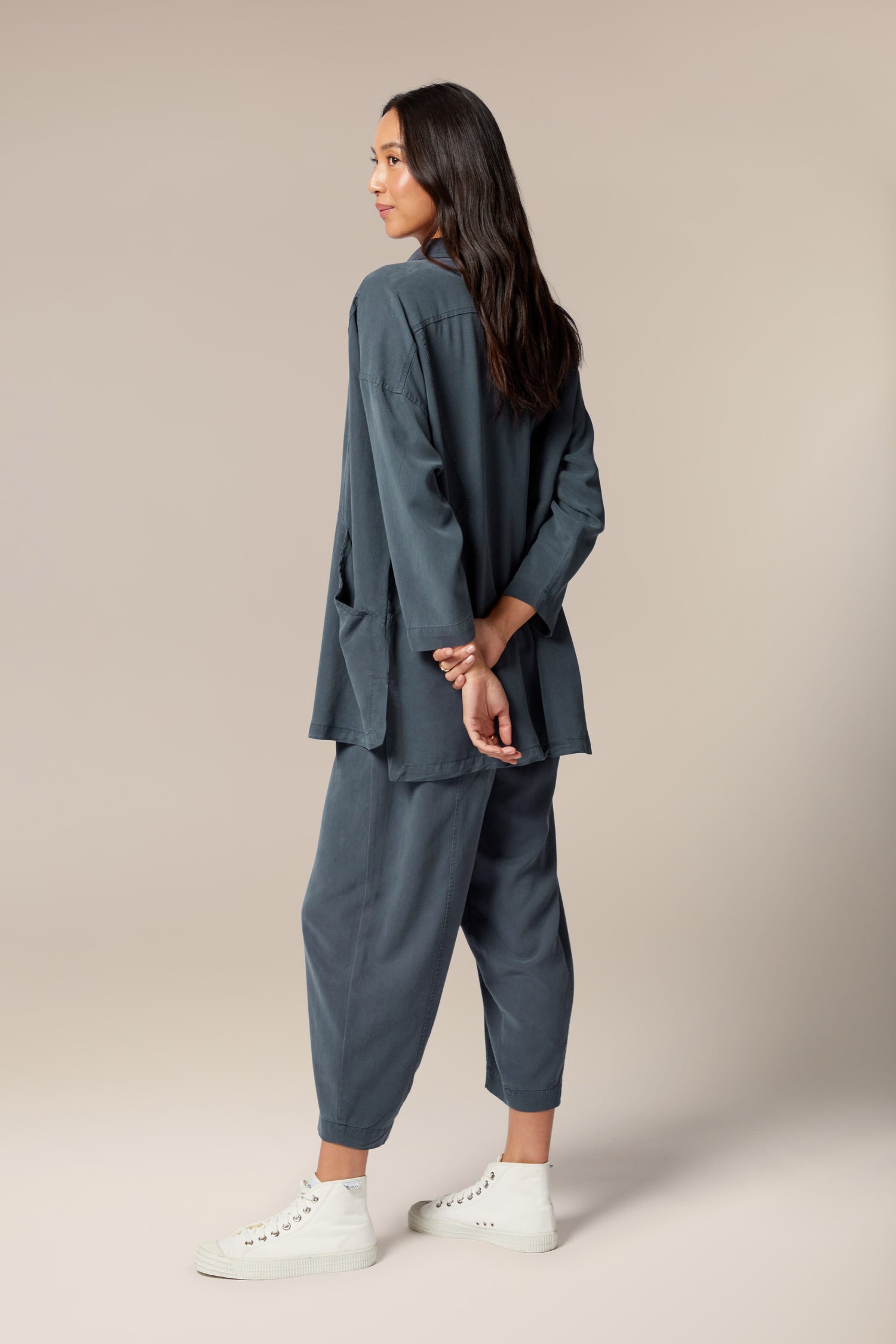 A model is wearing a blue twill shirt open with a collar, button front and pockets. It is paired with a white top underneath, black trouser, and white trainers.