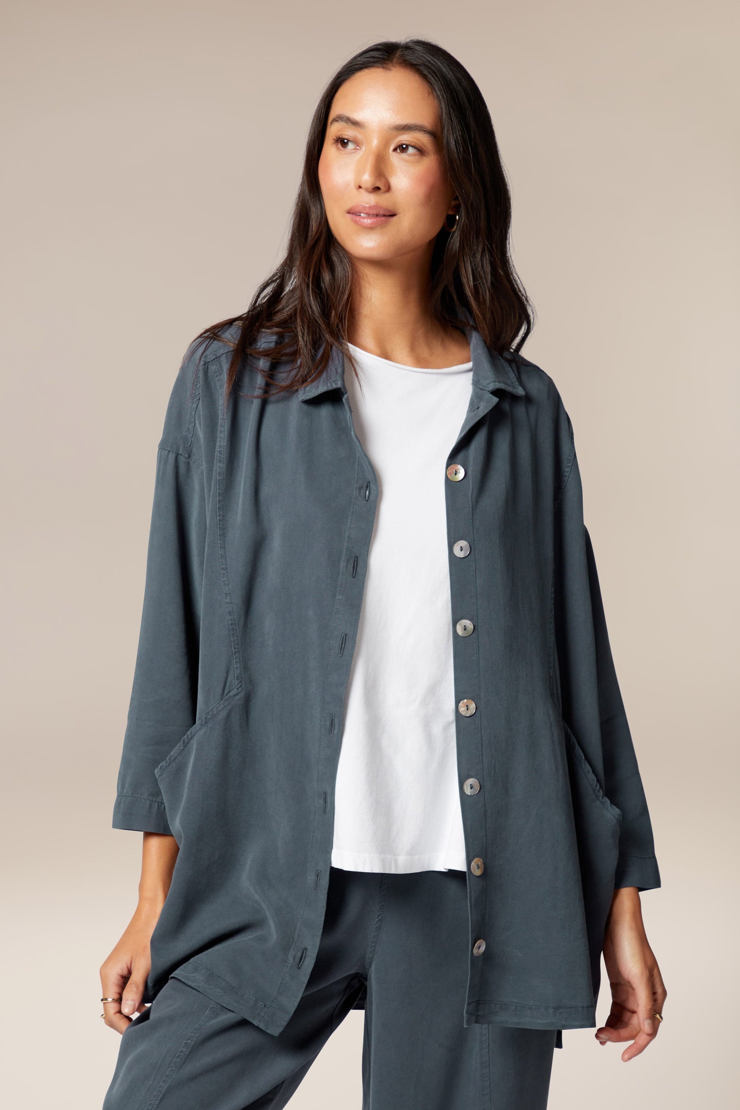 A model is wearing a blue twill shirt open with a collar, button front and pockets. It is paired with a white top underneath, black trouser, and white trainers.