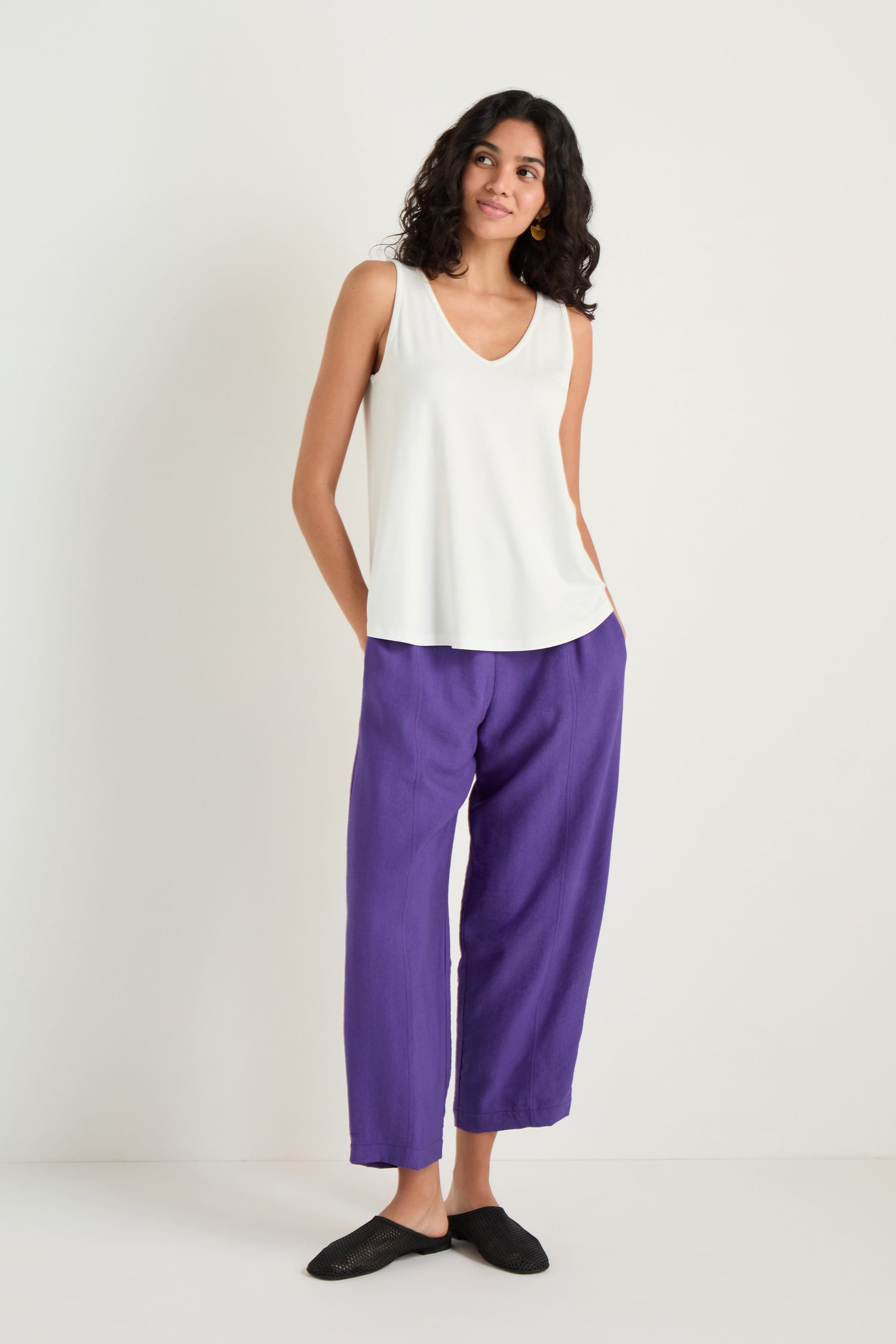 A woman stands against a white background wearing a Viscose Jersey Flare Vest, loose purple A-line pants, and black shoes, with her hands in her pockets.