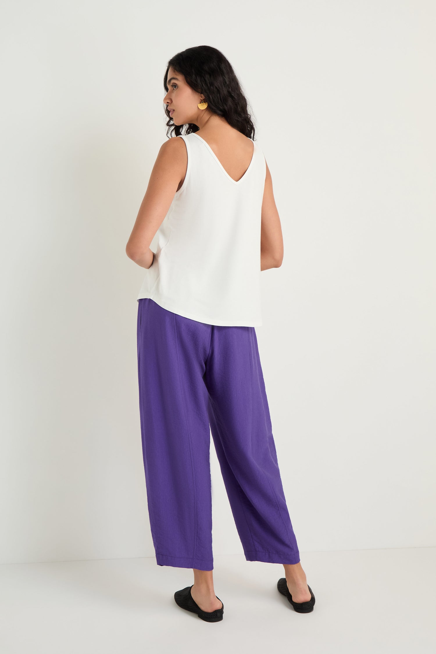 A woman faces away, wearing the Viscose Jersey Flare Vest—a sleeveless white top with a V-shaped back and A-line cut—paired with loose purple pants and black flats.