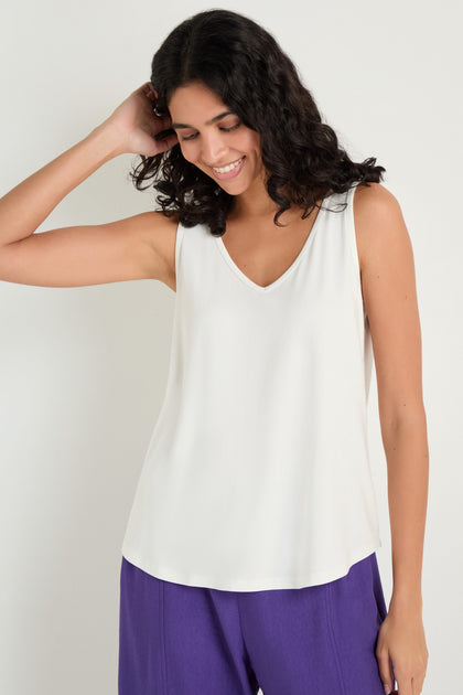A woman with medium-length curly hair wears the Viscose Jersey Flare Vest and purple pants, standing against a plain light background and smiling with one hand touching her hair.