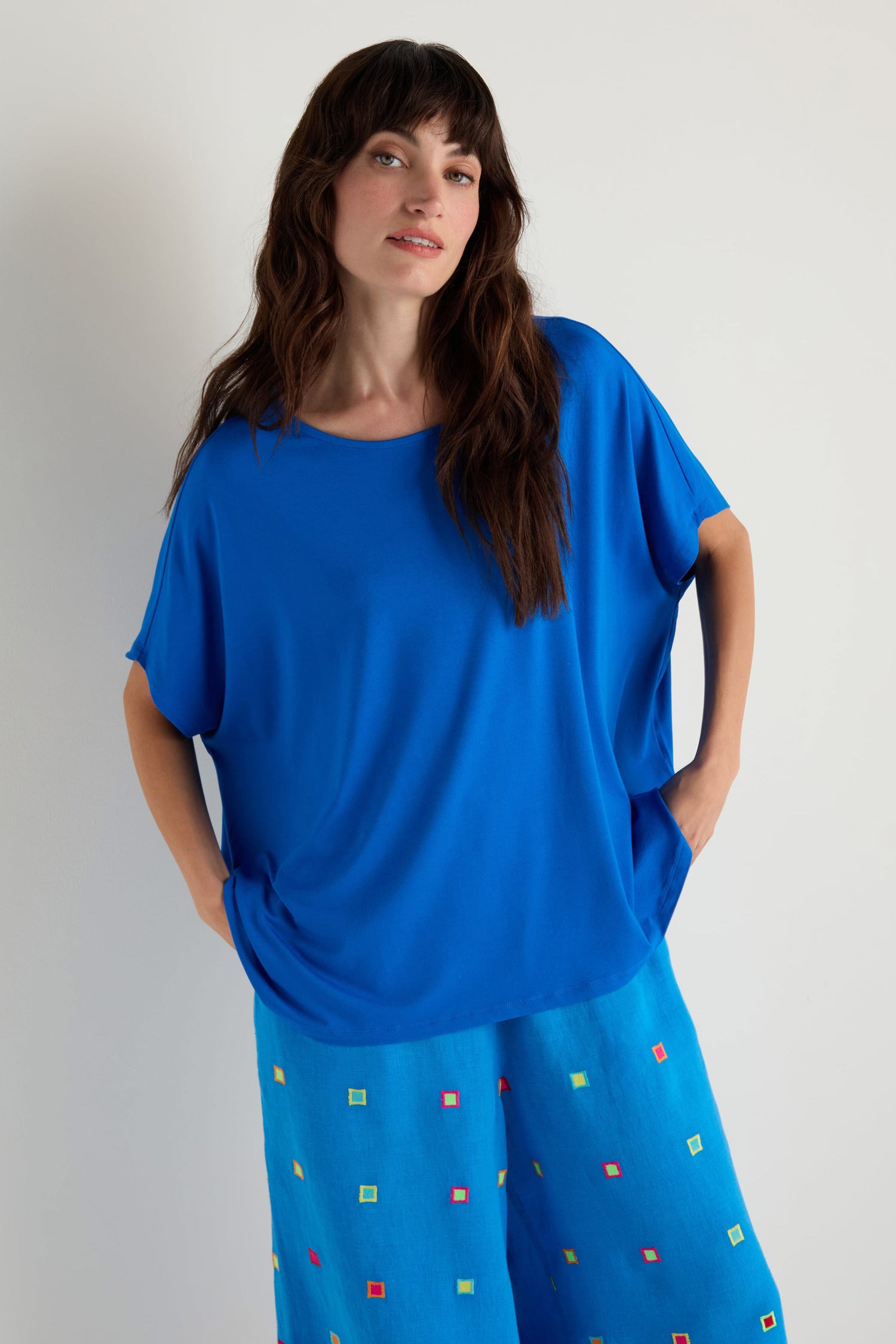 A woman with long brown hair wears the Viscose Jersey Boxy Top in bright blue and wide-leg blue pants with colorful square patterns, standing against a plain light background.