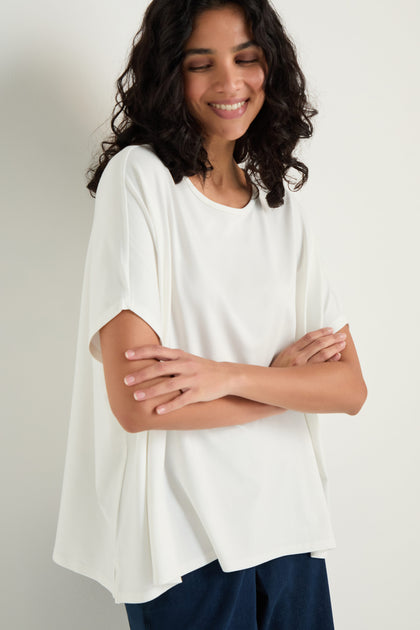 A woman with wavy hair stands against a light background, arms crossed and smiling slightly, wearing the Viscose Jersey Boxy Top in white with dark pants.