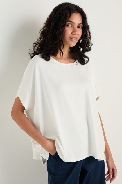 A woman with curly hair wears the Viscose Jersey Boxy Top and dark pants, standing against a plain light background with one hand in her pocket.