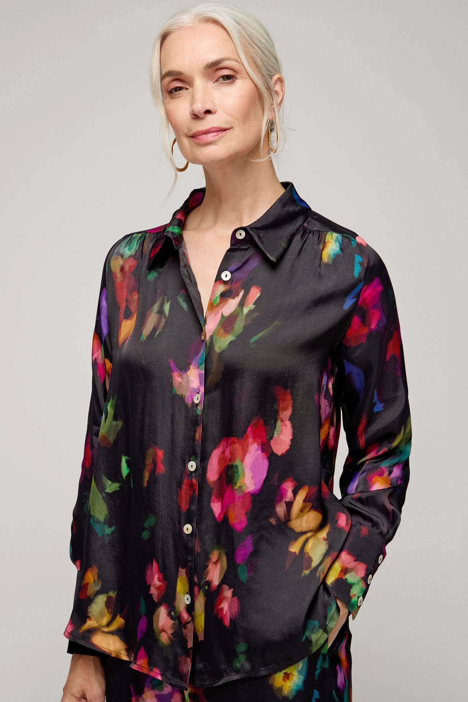 A woman with gray hair pulled back smiles slightly, wearing the Winter Floral Shirt—a black button-up with a painterly floral print—while standing against a neutral background.