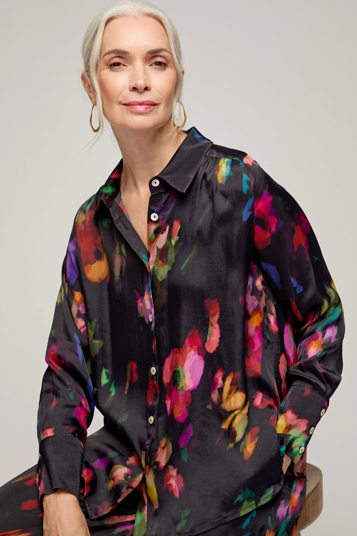 A woman with gray hair and large hoop earrings sits on a stool against a plain background, wearing the Winter Floral Shirt—a black button-up top featuring a painterly floral design.