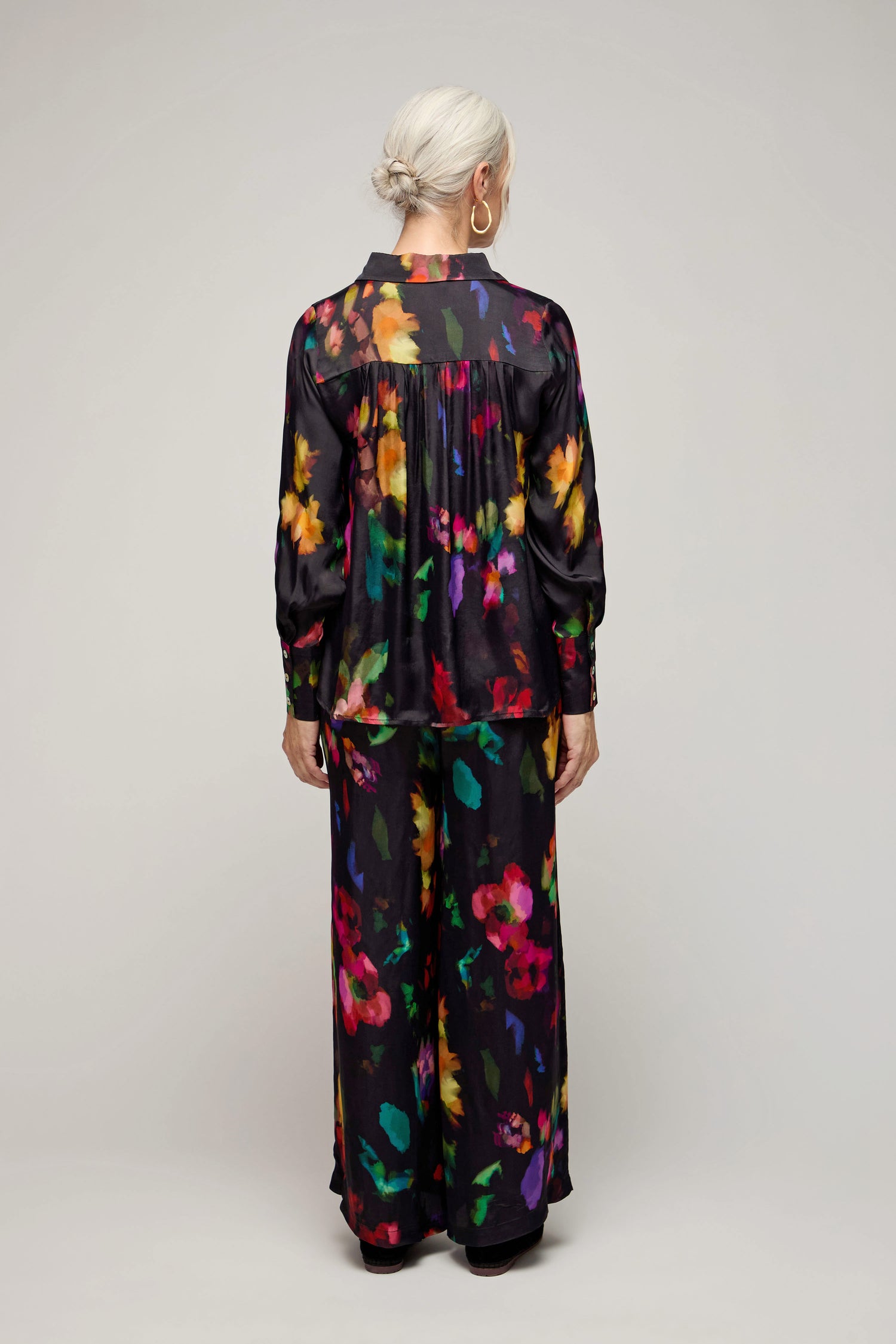 A person with light hair in a low bun wears the Winter Floral Shirt—long-sleeve, loose-fit, with a winter floral print—and wide-leg pants, shown from the back against a plain backdrop.