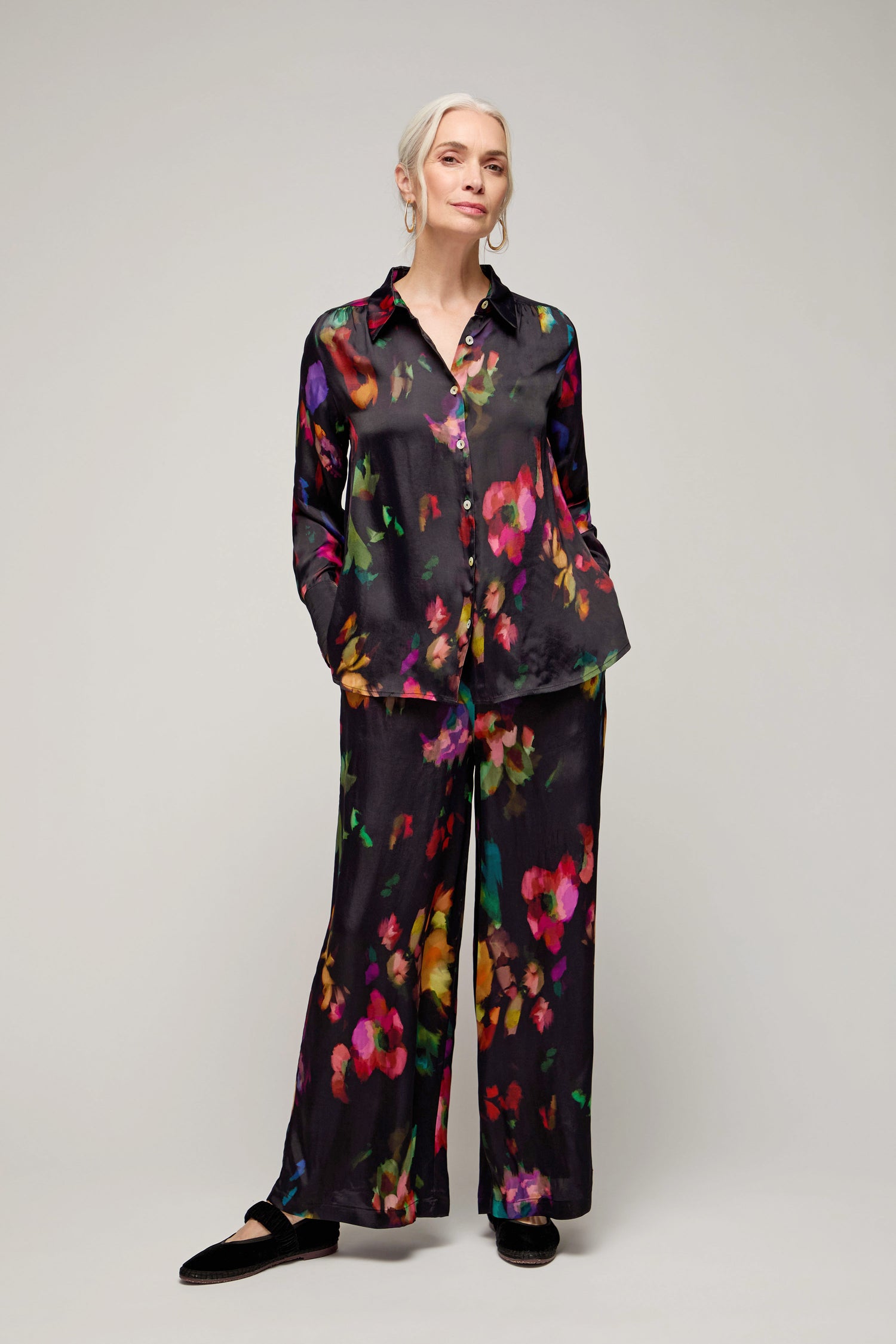 A woman stands against a plain background wearing the Winter Floral Shirt, a matching black outfit with a painterly floral print, hands in pockets, and black shoes.