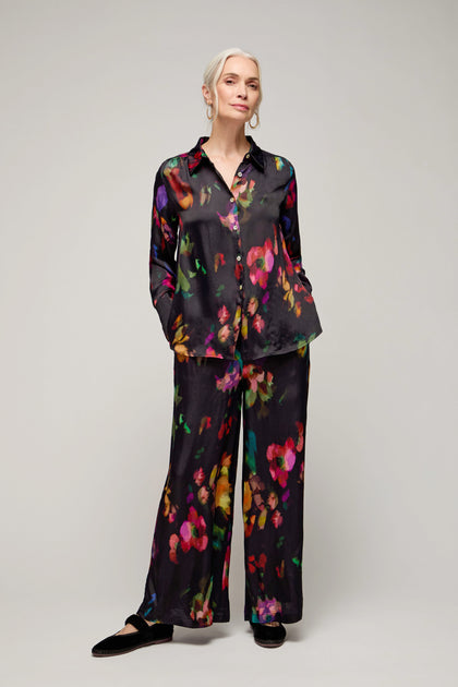 A woman stands against a plain background wearing the Winter Floral Shirt, a matching black outfit with a painterly floral print, hands in pockets, and black shoes.