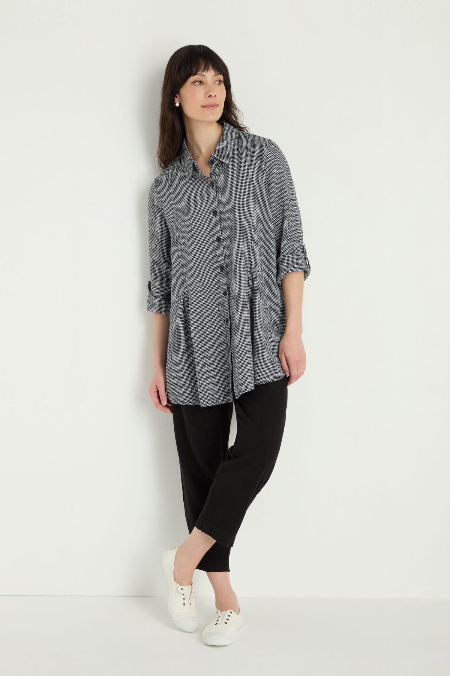 A woman stands against a white wall wearing the Micro Gingham Shirt in gray with long sleeves, paired with black pants and white sneakers. She gazes slightly to the side, arms relaxed at her sides.