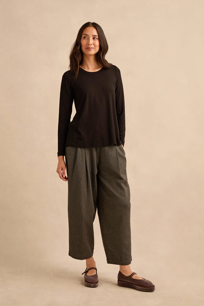 A woman stands against a beige background, wearing the Viscose Jersey Scoop Neck Top in black with long sleeves, paired with gray wide-leg pants and brown shoes, her arms relaxed and a slight smile on her face.