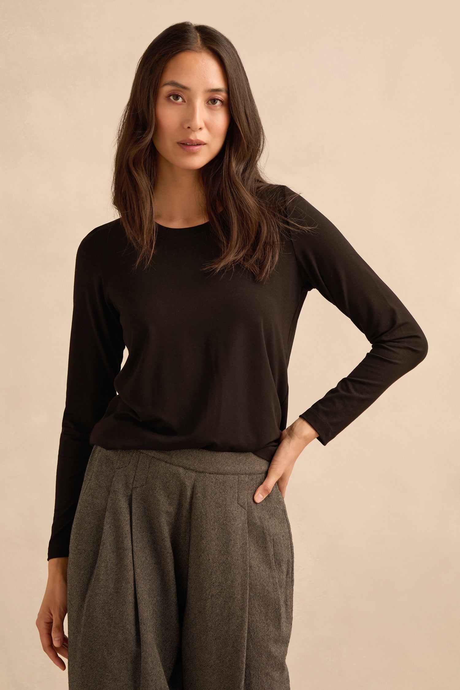 A woman wearing the Viscose Jersey Scoop Neck Top in black with gray pleated trousers stands against a neutral background, one hand on her hip.