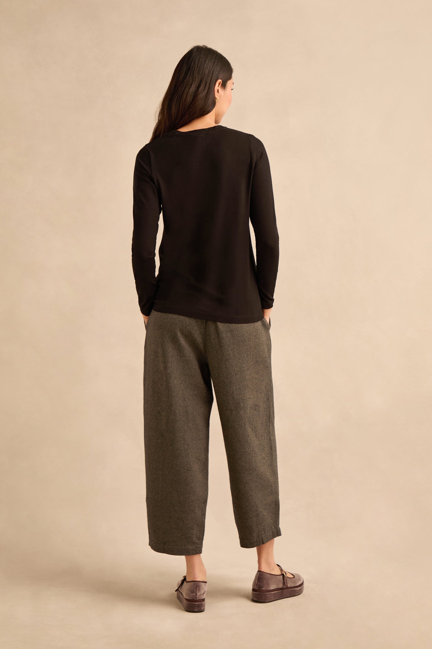 A woman stands with her back to the camera, wearing a black Viscose Jersey Scoop Neck Top with long sleeves, gray wide-leg pants, and brown shoes, hands in her pockets, against a beige background.