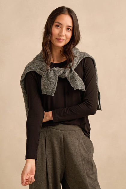 A woman stands against a neutral background, wearing the Viscose Jersey Scoop Neck Top in black with long sleeves, a grey sweater draped over her shoulders, and grey pleated pants.