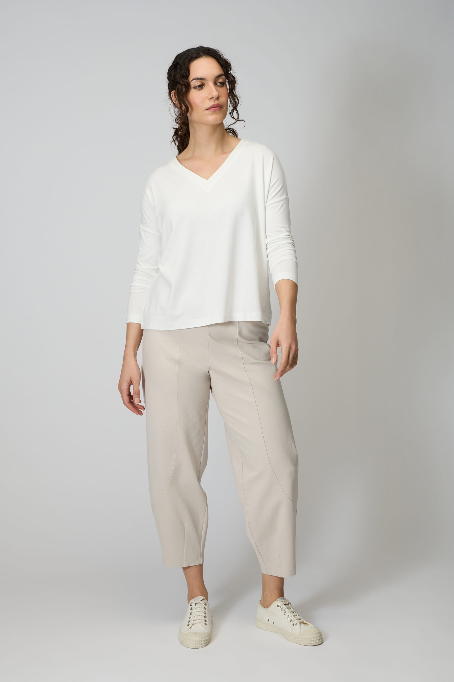A woman stands against a plain background, wearing a white Viscose Jersey V Neck Top with 3/4 sleeves, light beige cropped pants, and white sneakers.