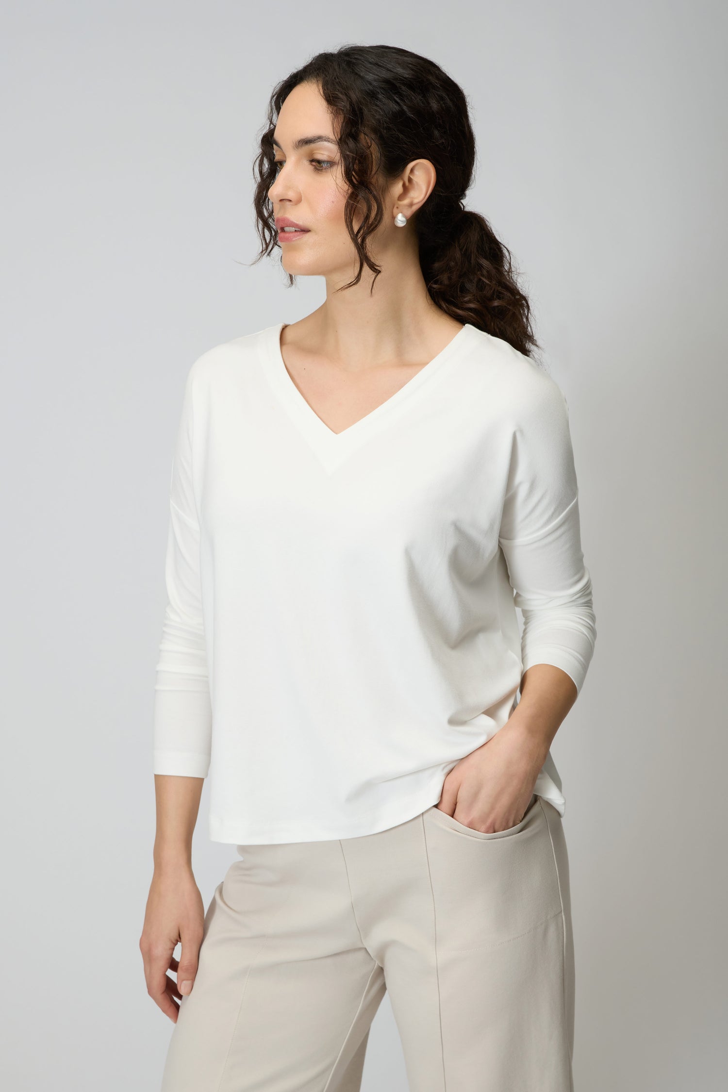 A woman wears the Viscose Jersey V Neck Top in white with 3/4 sleeves and beige pants, standing against a plain light background, looking to the side with one hand in her pocket.