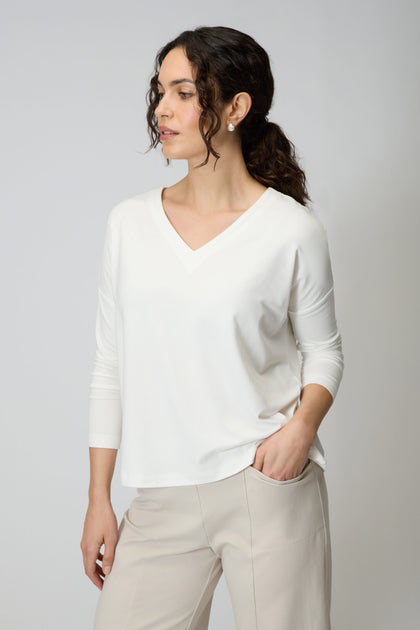 A woman wears the Viscose Jersey V Neck Top in white with 3/4 sleeves and beige pants, standing against a plain light background, looking to the side with one hand in her pocket.