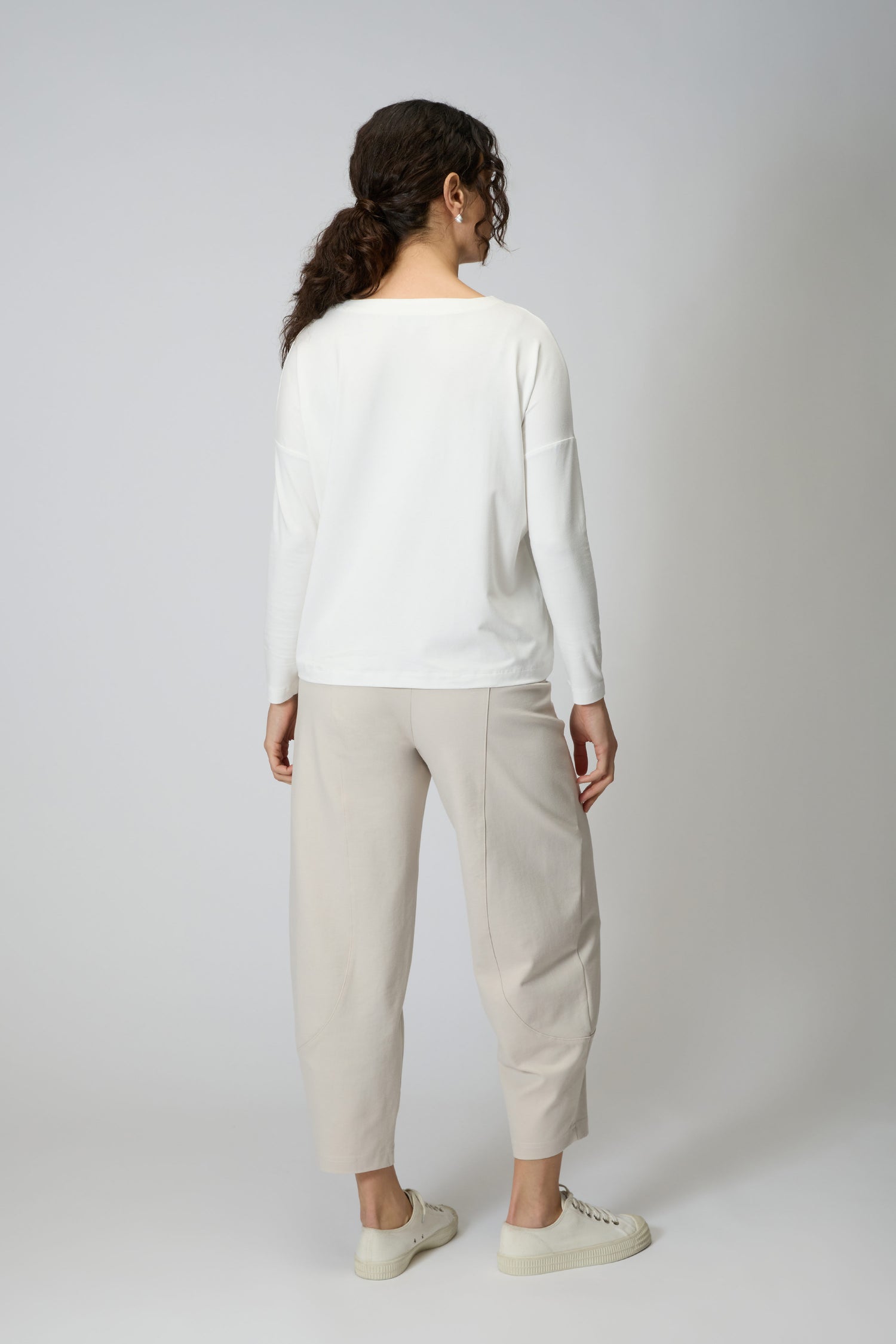 A person stands facing away against a plain light background, wearing the Viscose Jersey V Neck Top in white with 3/4 sleeves, paired with light beige pants and white sneakers.