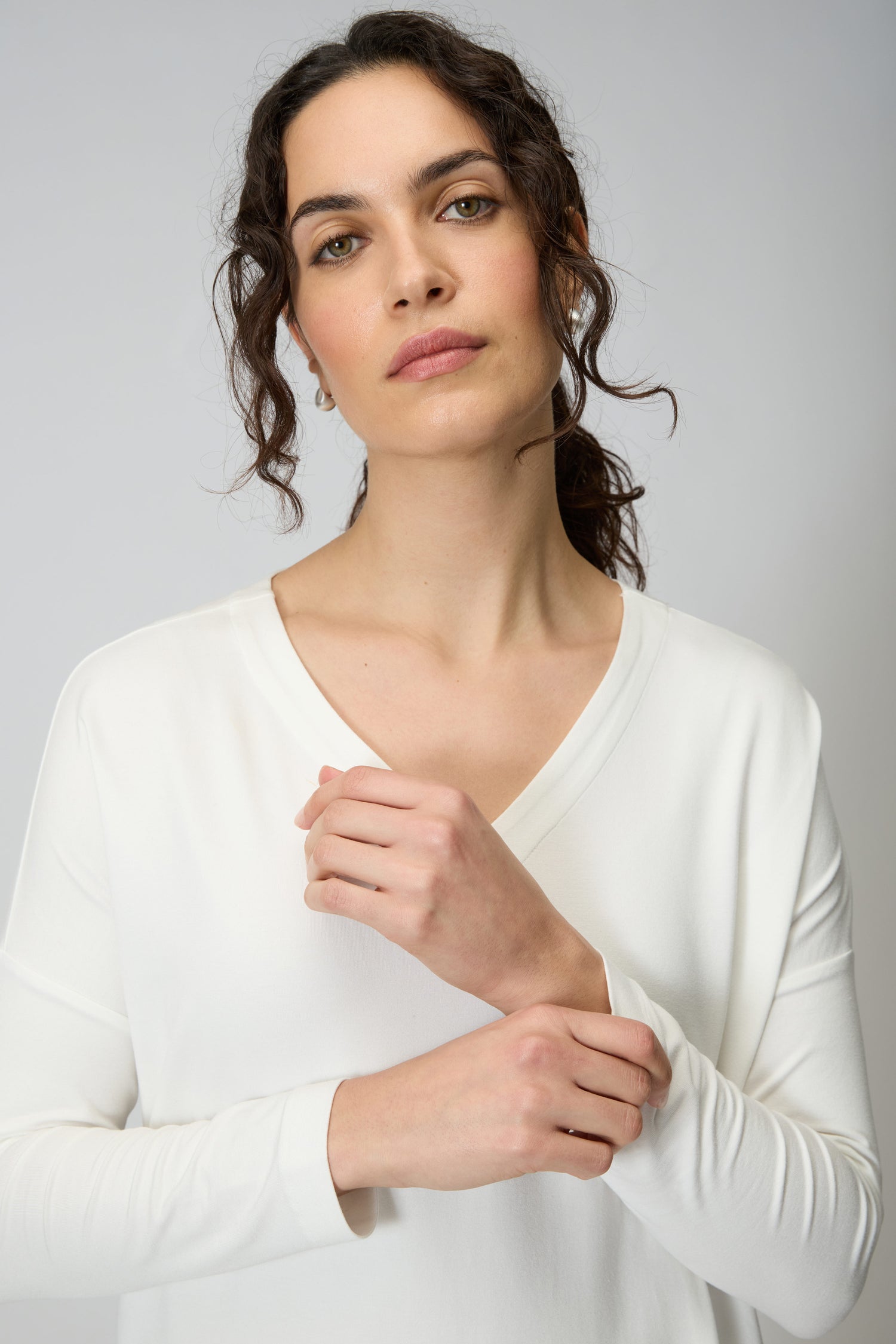 A woman with wavy brown hair in a ponytail wears the Viscose Jersey V Neck Top in white with 3/4 sleeves, calmly looking at the camera as she adjusts her sleeve, set against a plain light gray background.